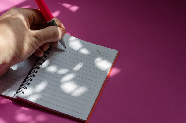 Світло-тіньова композиція лівої руки, що пише в блокноті на рожевому тлі. Чоловік середнього віку робить ноти з рожевою ручкою. Бізнес, повсякденна концепція. Вибірковий фокус
. - Фото, зображення