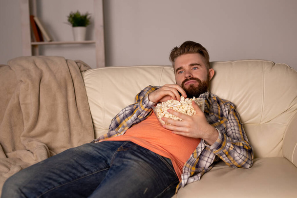 Hombre barbudo viendo películas o juegos deportivos TV comiendo palomitas de maíz en casa por la noche. Cine, campeonato y concepto de ocio. - Foto, imagen
