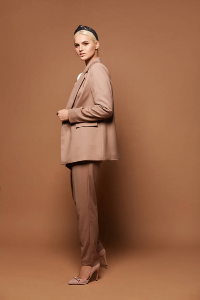Gorgeous woman with blond hair in a stylish suit posing against a beige background. Blond female model with perfect makeup wearing official outfit over beige background - Zdjęcie, obraz