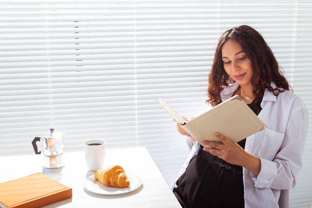 Side näkymä onnellinen raskaana oleva nainen lukee kirjaa, kun ottaa aamulla aamiaista kahvia ja croissants taustalla kaihtimet. Hyvää huomenta konsepti ja miellyttävä lounastauko - Valokuva, kuva