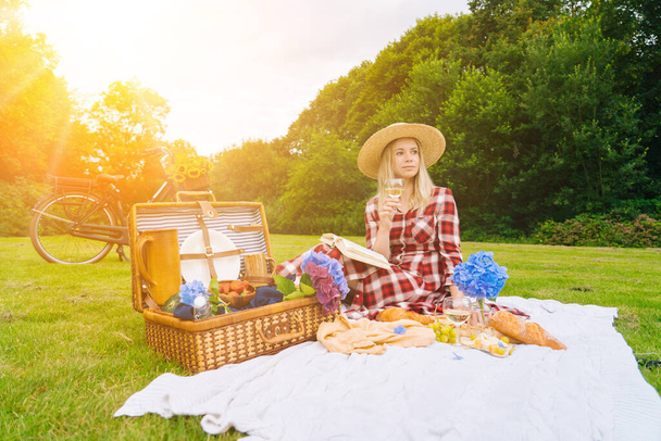 Girl in red checkered dress and hat sitting on white knit picnic blanket reading book and drinking wine. Summer picnic on sunny day with bread, fruit, bouquet hydrangea flowers. Selective focus. - Photo, Image