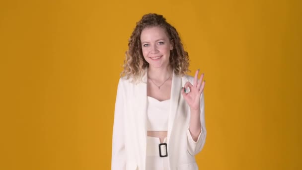 Positive young attractive girl with curly hair posing isolated on yellow studio background. Woman Looking approvingly at camera showing ok okay gesture, like sign positive something good. People lifes - Materiał filmowy, wideo