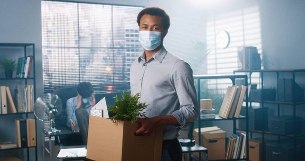 Portrait of young African American male worker in medical mask standing in cabinet holding carton box in hands with his stuff and looking at camera, fired from work, new employee at office - Foto, Imagen