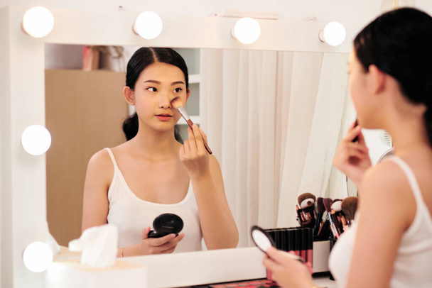 Mulher bonita olhando no espelho e aplicando cosméticos com uma escova grande - Foto, Imagem