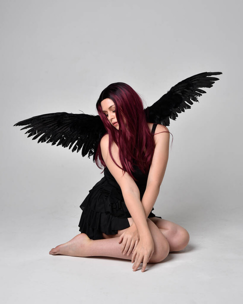 Retrato de cuerpo entero de una chica pelirroja con vestido negro y alas de ángel de plumas. Posar sentado sobre un fondo de estudio. - Foto, imagen