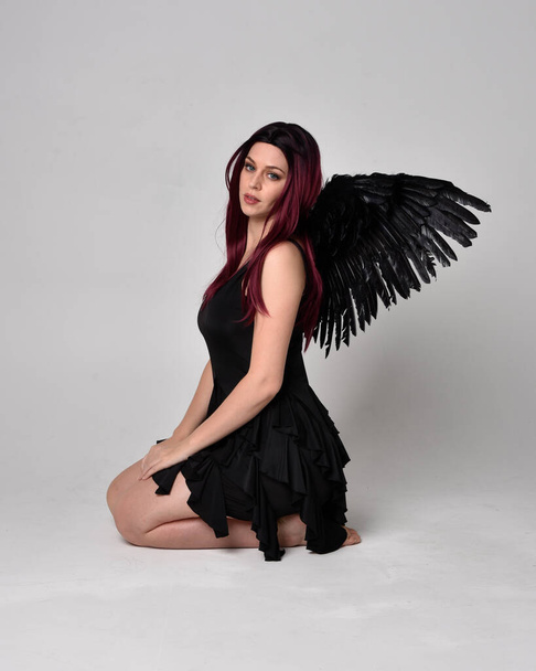 Portrait complet d'une fille aux cheveux roux portant une robe noire et des ailes d'ange en plumes. Pose assise sur un fond de studio. - Photo, image