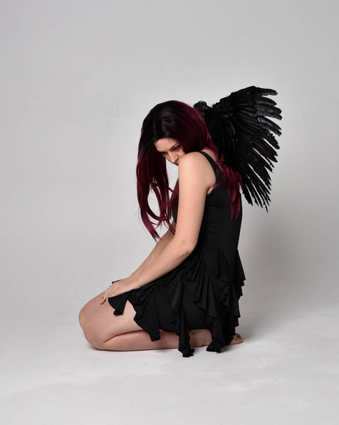 Портрет рудої волохатої дівчини в чорній сукні та ангельських крилах перо. Сидяча поза на фоні студії
. - Фото, зображення
