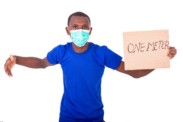 um jovem com uma camiseta azul em pé sobre um fundo branco usando uma máscara médica segurando um pôster com as mãos estendidas. - Foto, Imagem