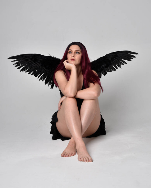 Portrait complet d'une fille aux cheveux roux portant une robe noire et des ailes d'ange en plumes. Pose assise sur un fond de studio. - Photo, image