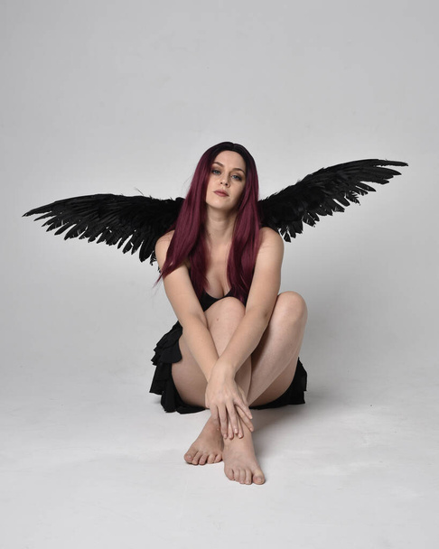 黒のドレスと羽の天使の翼を身に着けている赤い髪の少女の完全な長さの肖像画。スタジオを背景に座りポーズ. - 写真・画像