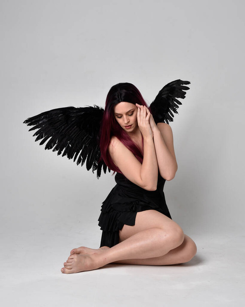 Полный портрет рыжеволосой девушки в черном платье и крыльях ангела-перышка. Сидящая поза на фоне студии. - Фото, изображение