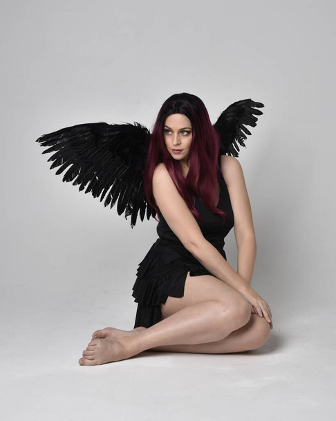Full length portrait of a red haired  girl wearing black dress and feather angel wings.  Sitting pose against a studio background. - Φωτογραφία, εικόνα