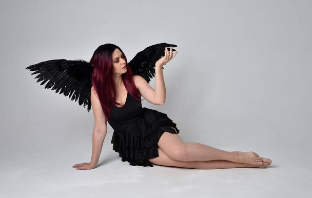 Retrato de comprimento total de uma menina de cabelos vermelhos usando vestido preto e asas de anjo de penas. Sentado pose contra um fundo de estúdio. - Foto, Imagem