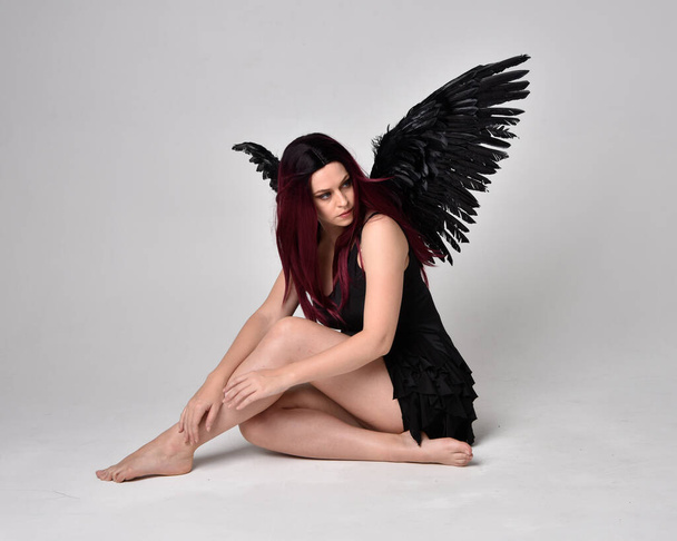 Full length portrait of a red haired  girl wearing black dress and feather angel wings.  Sitting pose against a studio background. - Photo, Image