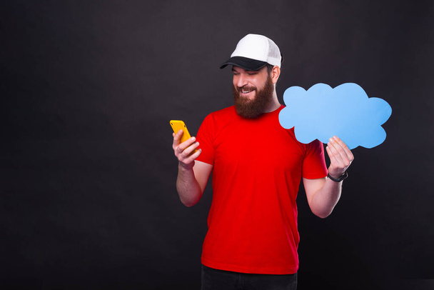 Fotografie vousatého muže v červeném tričku s použitím smartphonu a držící modrý oblak řeči. - Fotografie, Obrázek