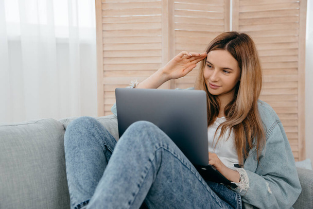 Szczęśliwa kobieta leżąca na kanapie w domu i patrząc na ekran komputera oglądanie webinar lub robi wideo czat przez kamerę internetową. - Zdjęcie, obraz