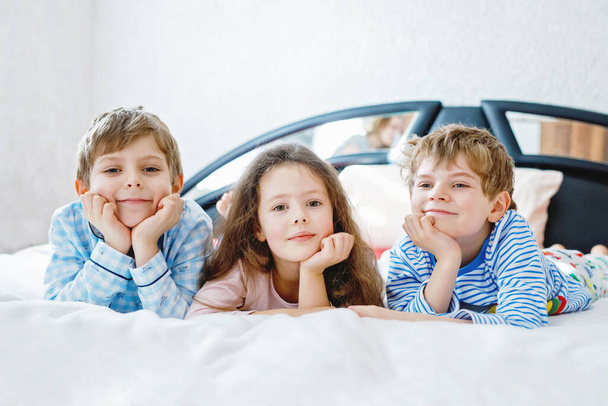 Three happy kids in pajamas celebrating pajama party. Preschool and school boys and girl in nightwear having fun together. Children playing together in bed. Making pillow fight, indoors at home. - Photo, Image
