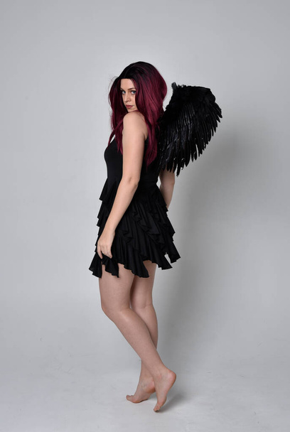 Portrait complet d'une fille aux cheveux roux portant une robe noire et des ailes d'ange en plumes. Position debout sur fond de studio. - Photo, image