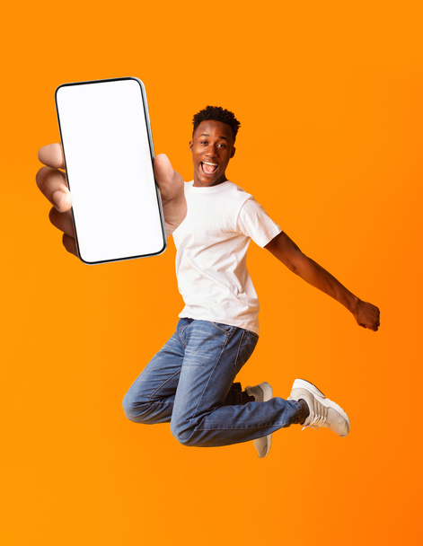 Überglücklicher schwarzer Mann springt und hält Smartphone mit weißem Bildschirm, Attrappe für Werbung oder App-Design - Foto, Bild