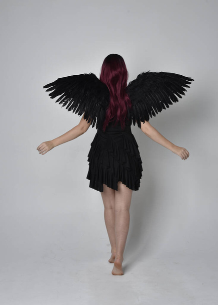 Full length portrait of a red haired  girl wearing black dress and feather angel wings.  Standing pose against a studio background. - Photo, Image