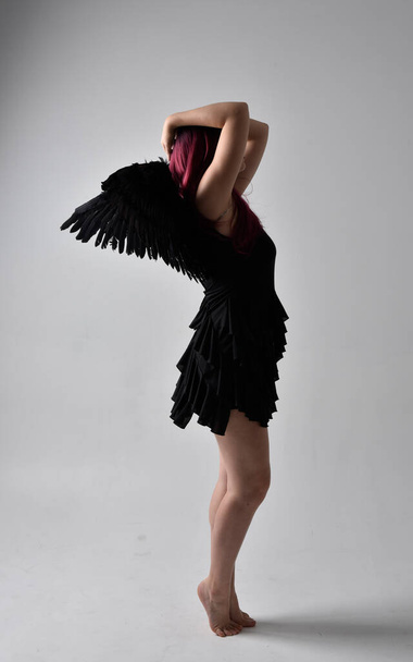 Retrato de comprimento total de uma menina de cabelos vermelhos usando vestido preto e asas de anjo de penas. pose de pé contra um fundo de estúdio. - Foto, Imagem