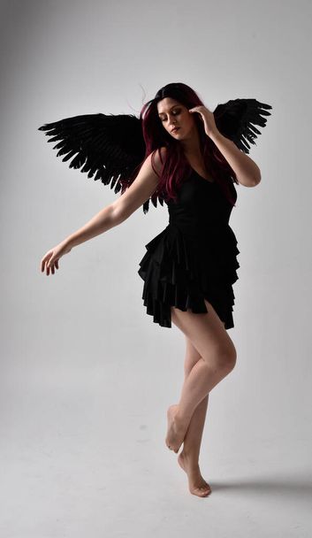 Full length portrait of a red haired  girl wearing black dress and feather angel wings.  Standing pose against a studio background. - Foto, afbeelding