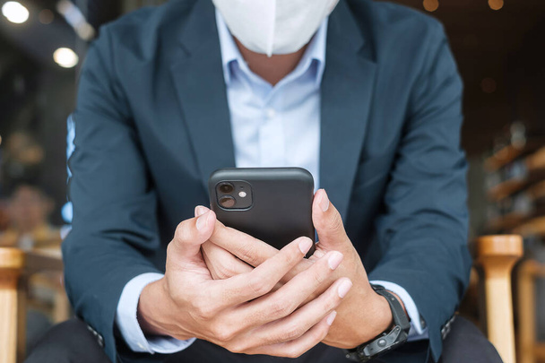 fiatal üzletember öltönyben sebészeti maszkot visel és okostelefont használ, férfi érintőképernyős mobiltelefont gépel az irodában vagy a kávézóban. Covid-19 világjárvány, technológia és új normális koncepció - Fotó, kép