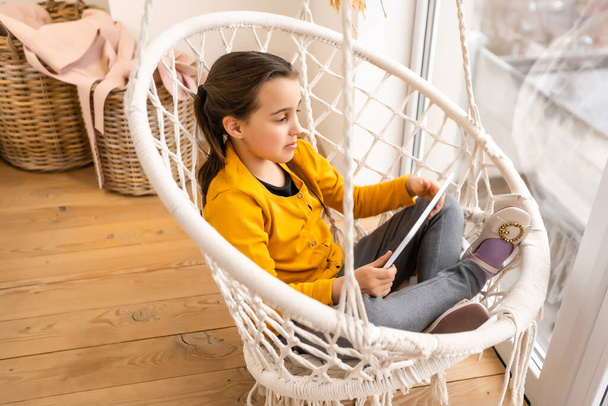 Улыбается маленькая девочка, лежащая на удобном диване, наслаждаясь игрой в онлайн на цифровом планшетном компьютере. В дополнение к технологиям, счастливый маленький ребенок с помощью смешных приложений, веб-серфинг информации - Фото, изображение