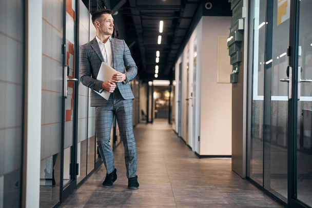 Handsome young man with laptop standing in office corridor - Photo, image