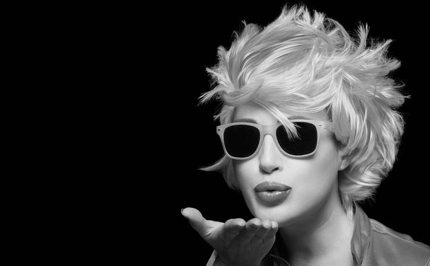 Stylish young woman with trendy modern sunglasses or shades and matching pink wig blowing across the palm of her hand sending a kiss with pouting lips. Monochrome closeup portrait isolated on black background - Photo, image