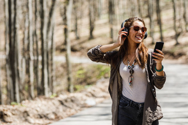 Φωτογραφία μιας χαμογελαστής νεαρής γυναίκας που χρησιμοποιεί το smartphone της και ακούει μουσική ενώ περπατάει στη φύση. - Φωτογραφία, εικόνα