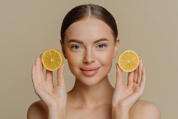 Headshot of beautiful brunette woman holds juicy lemon slices has healthy shiny skin gets vitamins from citrus stands bare shoulders indoor against beige background. Facial treatment concept - Photo, Image