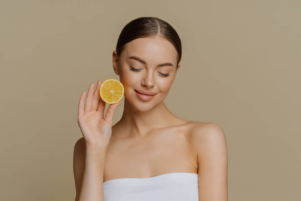 Portrait de belle brune jeune femme concentrée vers le bas détient tranche de citron frais recommande des produits de beauté naturels préfère un traitement facial biologique naturel enveloppé dans une serviette de bain pose à l'intérieur - Photo, image
