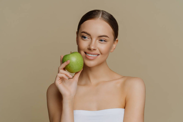 Nachdenklich charmante Europäerin hält Apfel in der Nähe Gesicht lächelt sanft hat weiße perfekte Zähne gesunde saubere Haut in Duschtuch umwickelt steht mit nackten Schultern vor braunem Hintergrund. - Foto, Bild