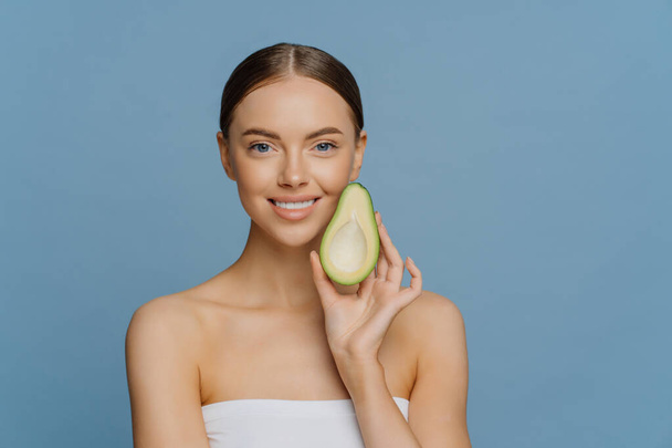Horizontal shot of happy brunette woman holds half of avocado near face uses anti aging organic recipes stands refreshed indoor wrapped in towel isolated over blue background. Healthy diet concept - Photo, Image