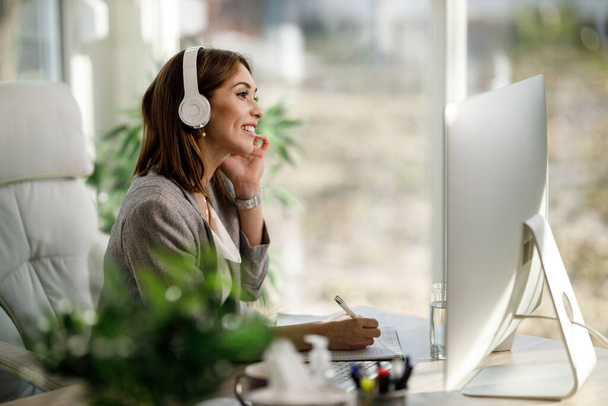 Μια ελκυστική επιχειρηματίας κάθεται μόνη της στο γραφείο του σπιτιού της με ακουστικά και εργάζονται σε υπολογιστή. - Φωτογραφία, εικόνα