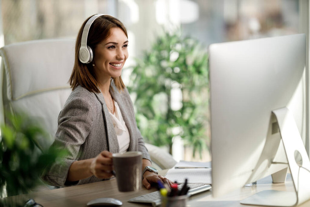 Uma mulher de negócios atraente sentada sozinha em seu escritório com fones de ouvido e trabalhando no computador. - Foto, Imagem