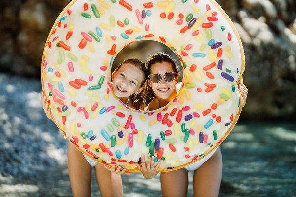 Eine schöne Teenagerin und ihre junge Mutter haben Spaß und schauen durch aufblasbare Gummikreise am Strand.  - Foto, Bild