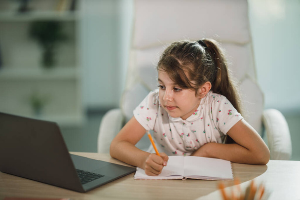 Ένα χαριτωμένο επιμελές κοριτσάκι που χρησιμοποιεί το laptop της για να κάνει ένα online μάθημα στο σπίτι. - Φωτογραφία, εικόνα