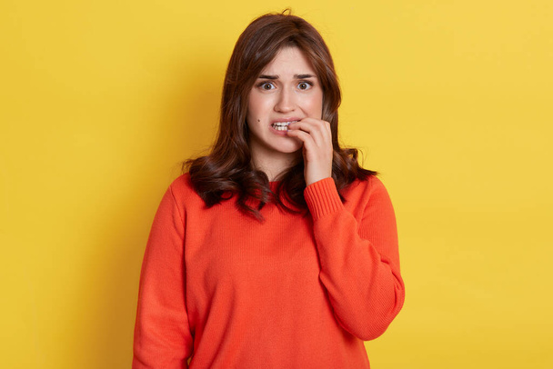 Nervous dark haired woman looking directly at camera with frightened expression, biting finger nails, being afraid of something, looks worried, posing isolated over yellow background. - Foto, imagen