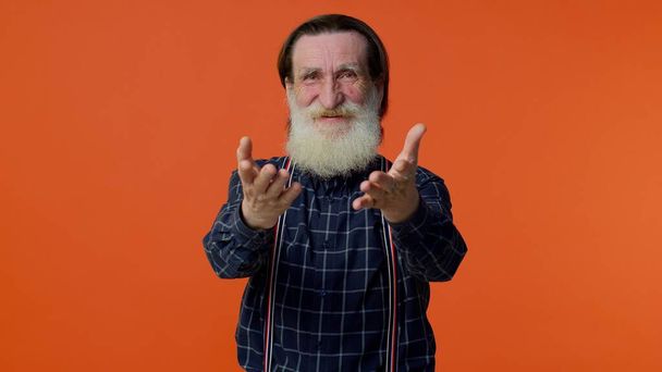 Senior smiling old man with gray-haired looking at camera, fixing beard posing on orange background - Фото, изображение