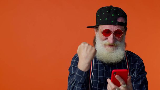 Літній бородатий чоловік використовує мобільний телефон, який переглядає онлайн кажучи: "Так, великі новини перемоги роблять жест переможця"
 - Фото, зображення