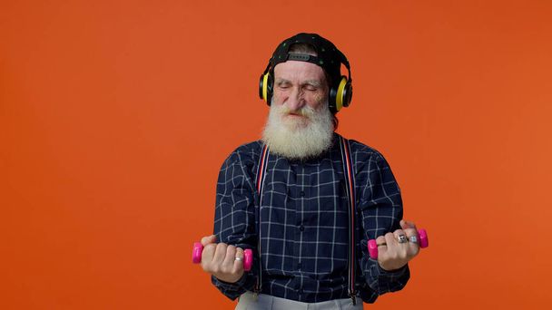 Anciano barbudo escuchando música a través de auriculares, haciendo ejercicio, levantando pesas rosadas, atención médica - Foto, Imagen