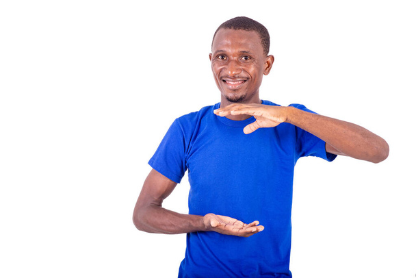 un jeune homme en t-shirt bleu debout sur un fond blanc montrant un grand signe et regardant la caméra, souriant. - Photo, image