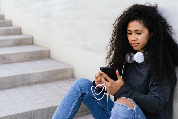 Hermosa chica afro de pelo rizado sentado en la calle utilizando el teléfono móvil con auriculares - Foto, imagen