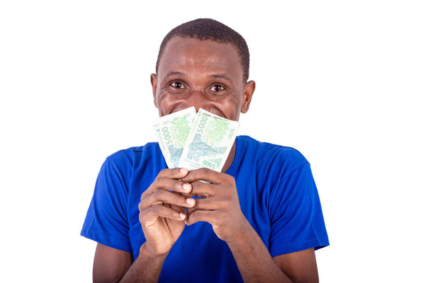 młody mężczyzna w niebieskiej koszulce stojący na białym tle z banknotami na ustach i patrzący w aparat uśmiechnięty. - Zdjęcie, obraz