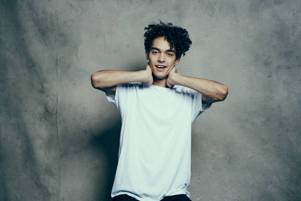 Cheerful guy with curly hair gestures with his hands near his face on a gray fabric background in a white t-shirt - Foto, afbeelding