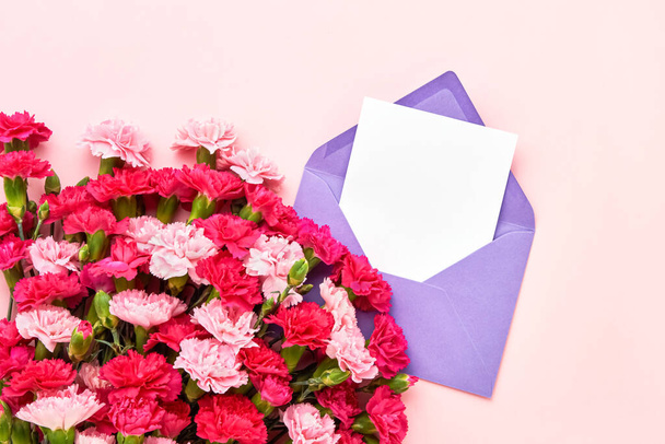 Rosafarbene Nelkenblüten und lila Umschlag auf rosa Pastellgrund. Geburtstag, Muttertag, Junggesellenabschied, Hochzeitskonzept. Kopierraum, Draufsicht. Grußkarte - Foto, Bild