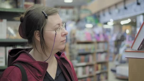 Serieuze blanke vrouw met bril op ketting koopt een boek in boekwinkel - Video