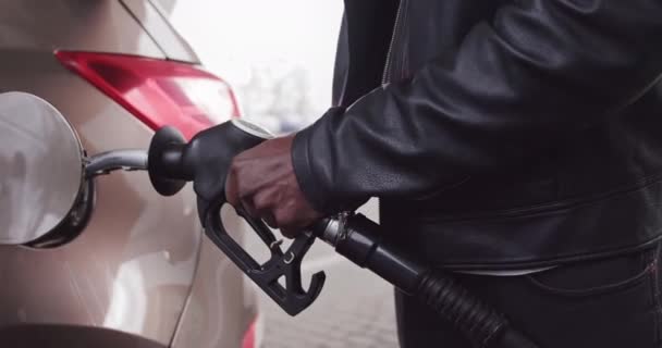 Recortado de cerca la imagen de empresario africano irreconocible en ropa casual, rholding en pistola de llenado a mano y repostando su coche de lujo con gasolina en la gasolinera - Metraje, vídeo